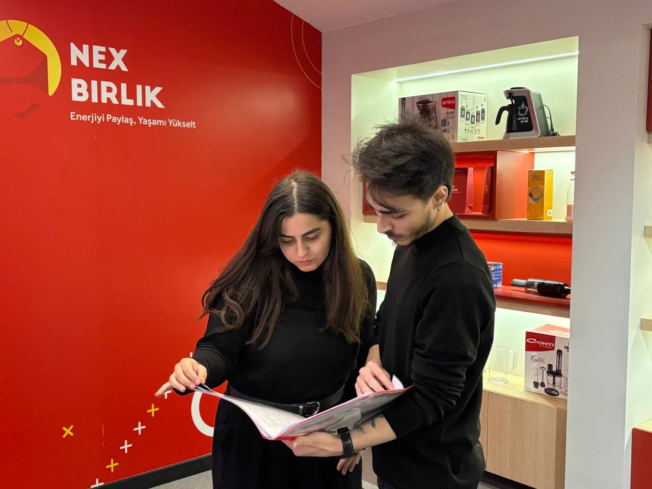 Qırmızı brend divarı önündə materiala baxan iki nəfər
